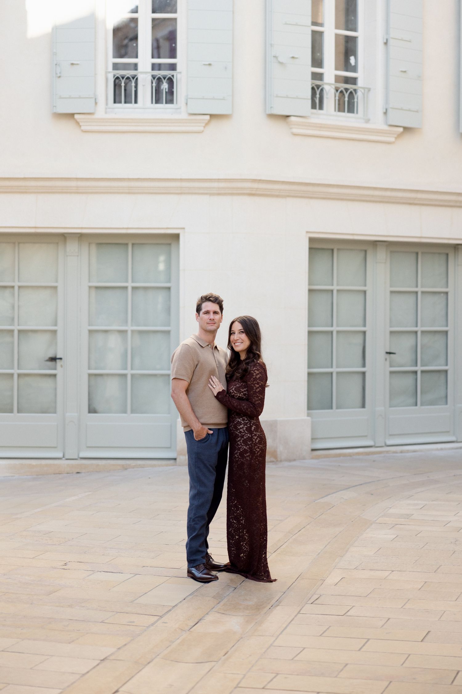 A couple stands together outside a classic French building with pale walls and light blue shuttered windows. Engagement session Paris