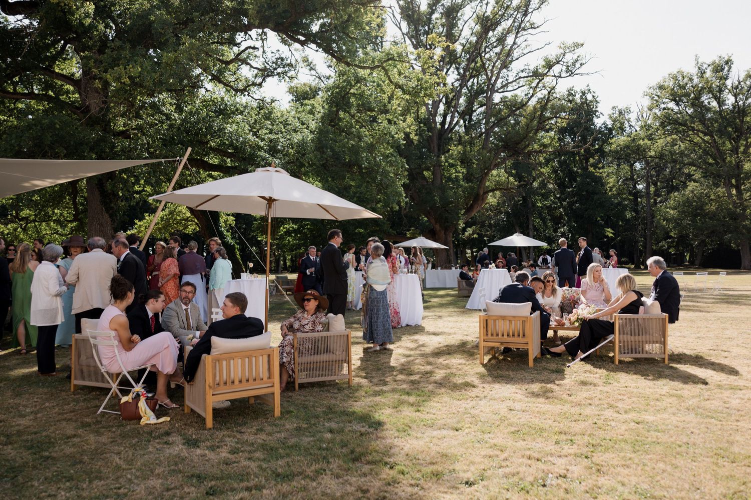 Outdoor garden party setup with wooden benches and white tents arranged under shady trees on a sunny afternoon.