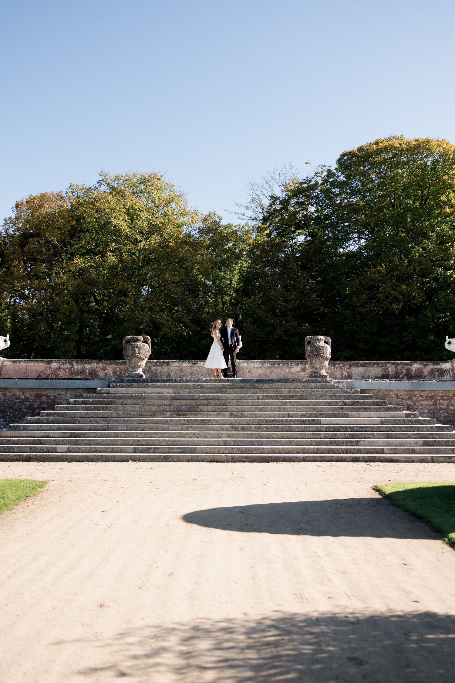 Bride and groom standing together atop grand stone staircase flanked by urns in formal garden.