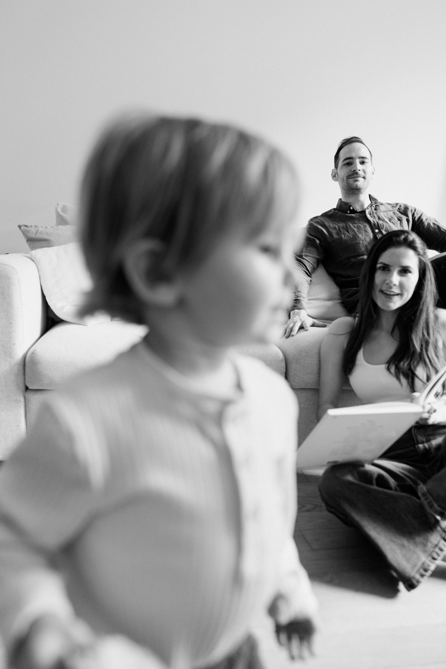 Black and white photo of happy parents relaxing on sofa watching their toddler child play on the floor.