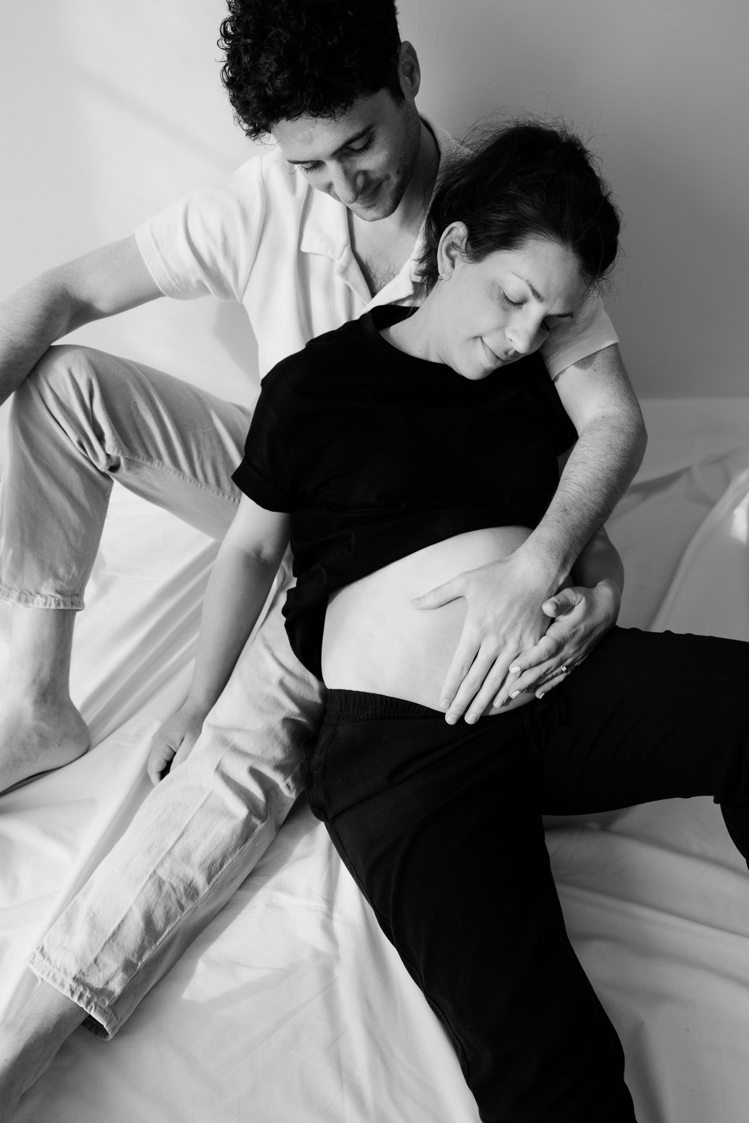 Black and white photo of a couple tenderly cradling a pregnant belly while sitting together on a bed.