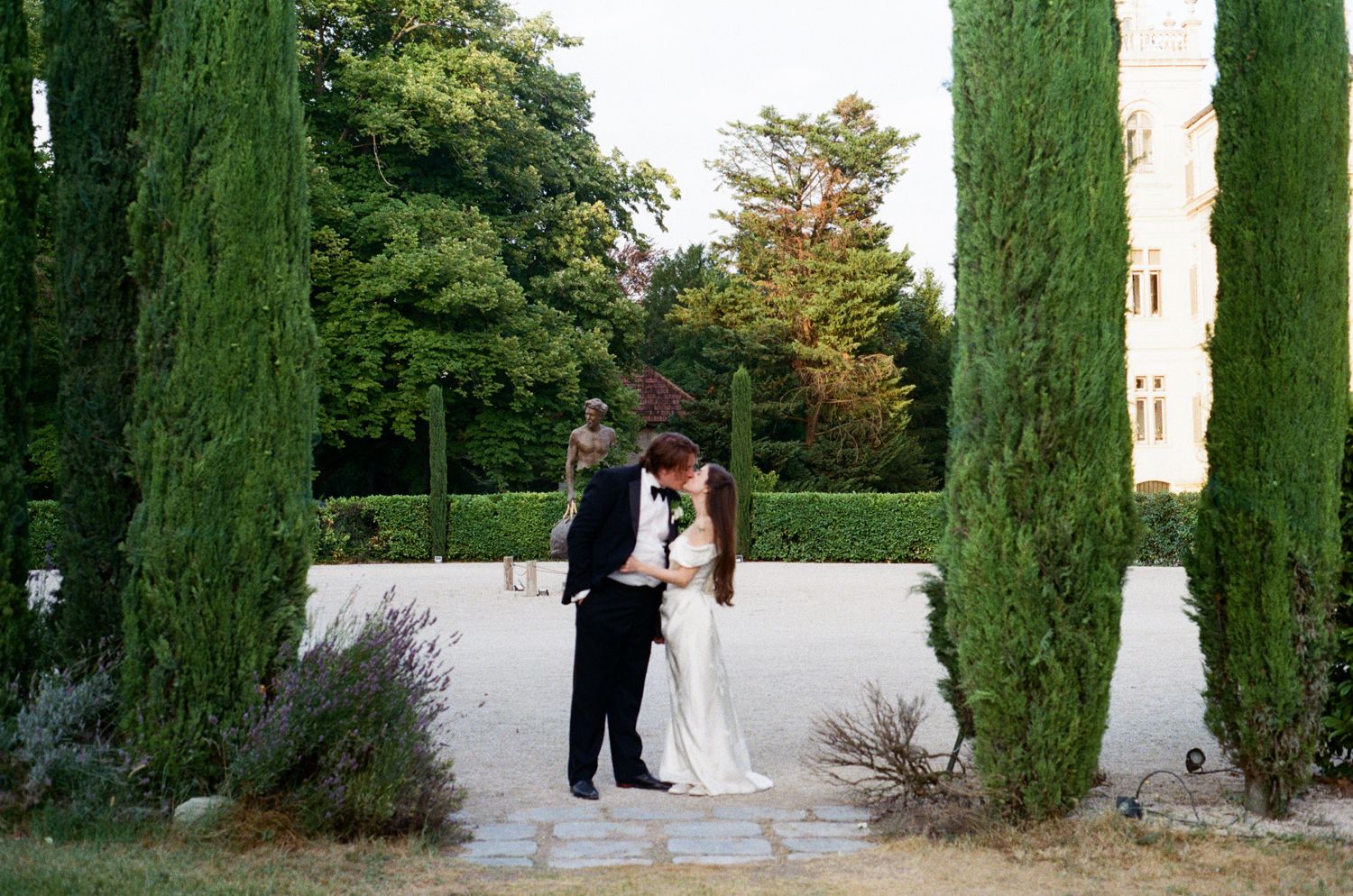 Couple standing in a garden pathway lined with tall cypress trees captured in a circular frame view.