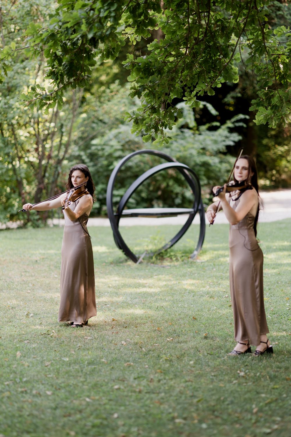 A figure in a brown dress stands in different poses on grassy lawn with large metal sculptures in the background.