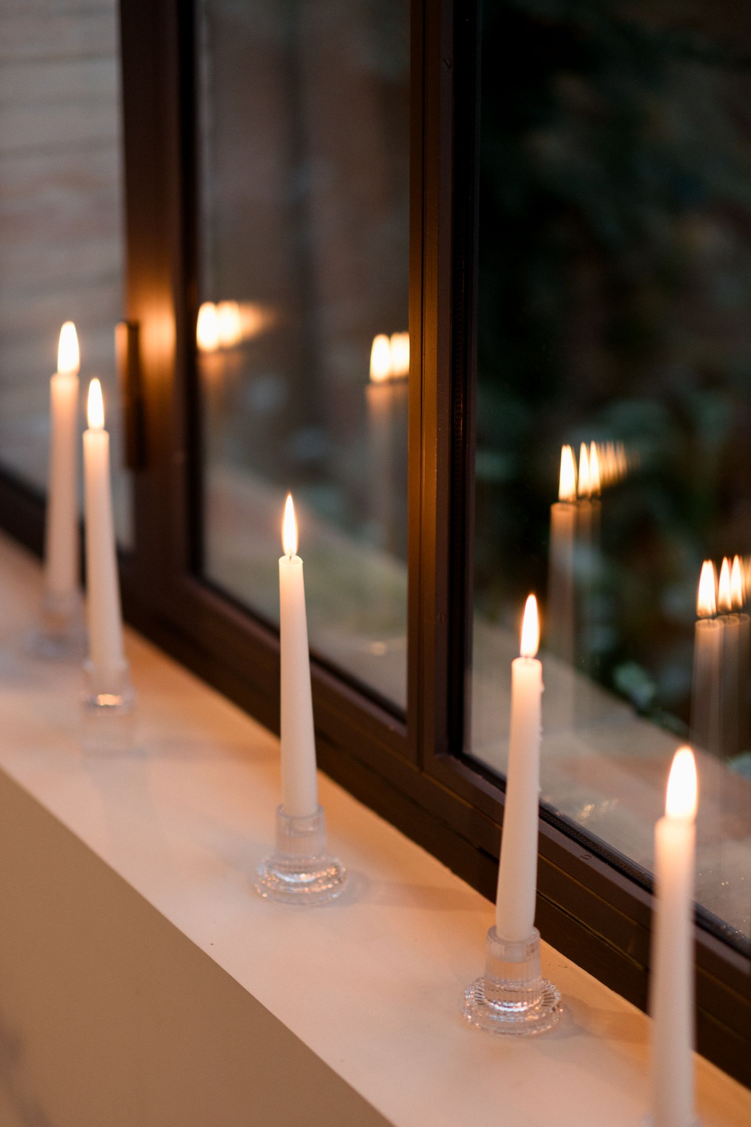 White taper candles in glass holders line a windowsill, creating a warm and cozy atmosphere with their glowing flames.