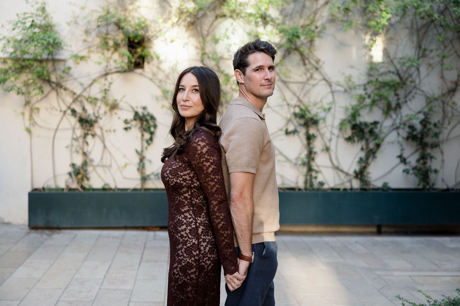Couple stands back-to-back holding hands against white wall with climbing vines, woman wearing brown lace dress. Engagement session France