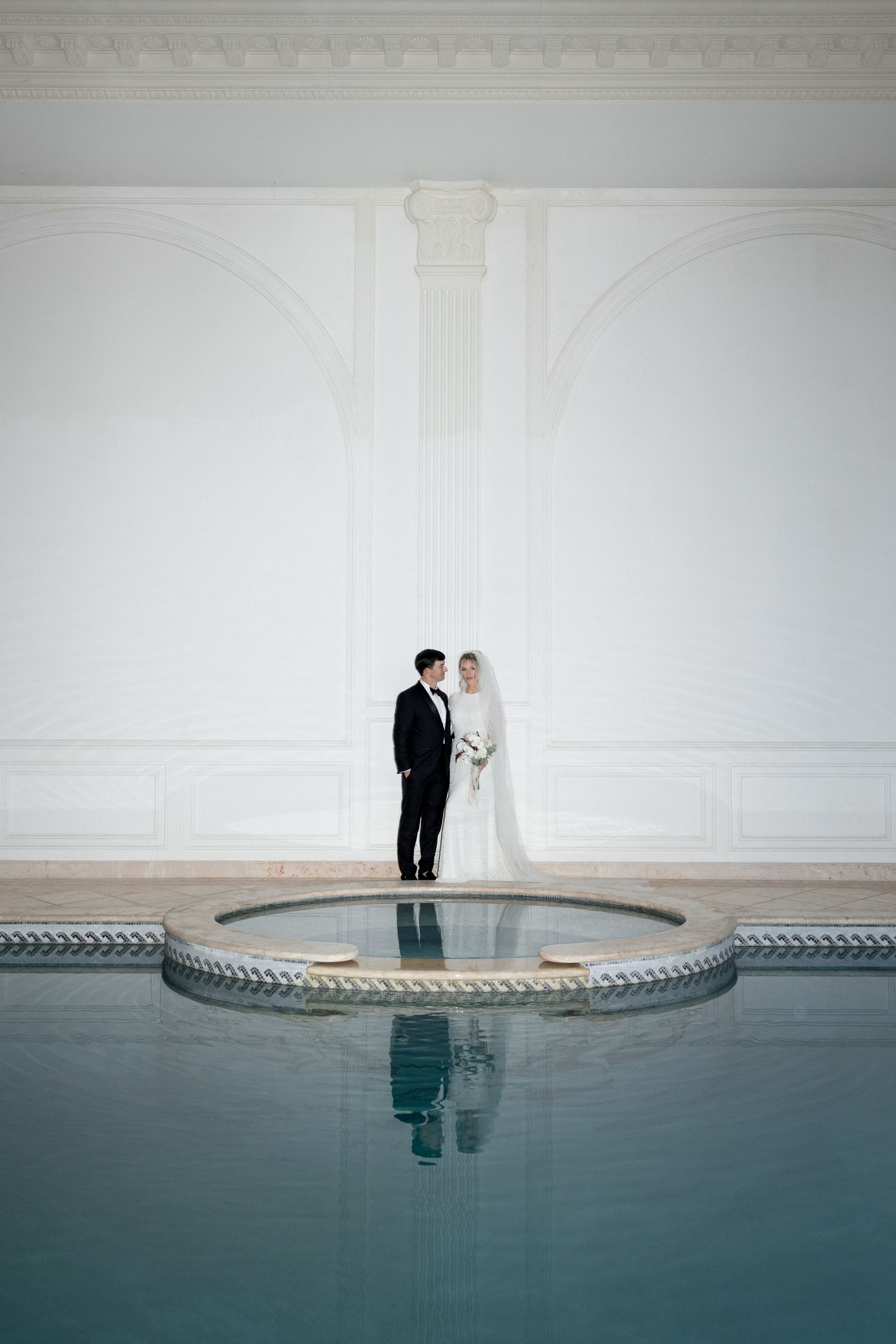 A couple stands together on a white platform overlooking a serene blue pool in a minimalist wedding setting. editorial wedding photographer