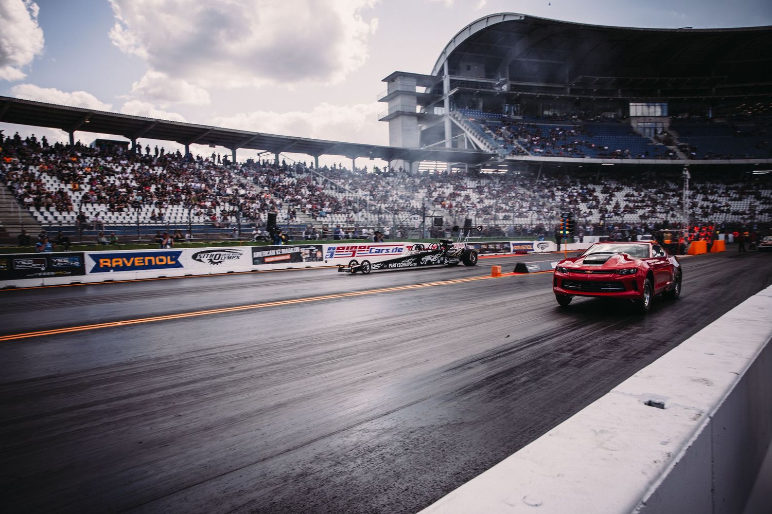 Side-by-side racing cars speed down a drag strip with spectator stands visible in the background.