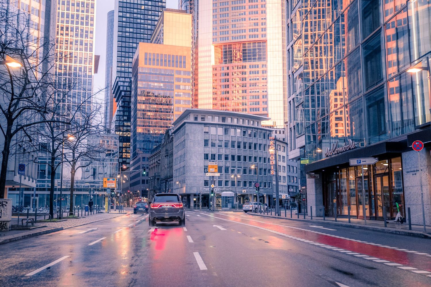 A moody urban cityscape at dusk with wet streets, red taillights, and golden reflections on modern skyscrapers.
