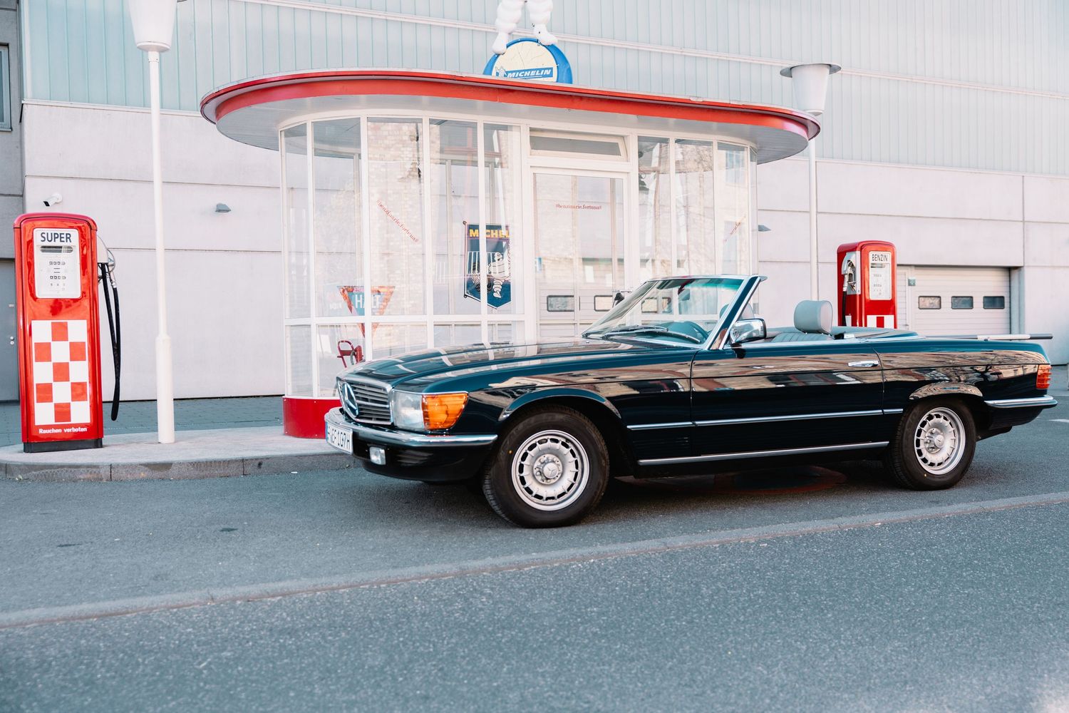 Classic black Mercedes convertible parked at a vintage-style gas station with red and white retro fuel pumps.