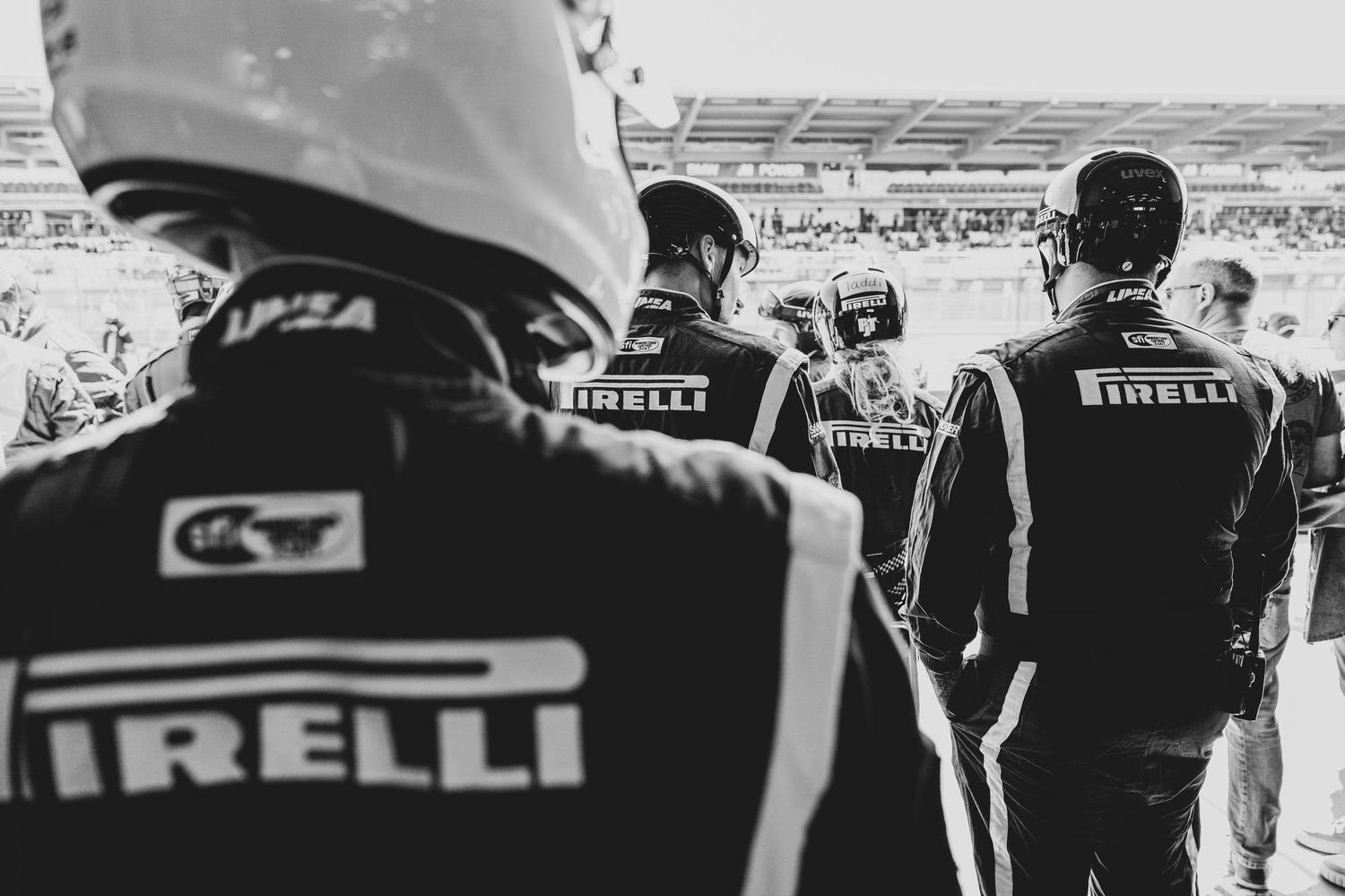 Pirelli team members in branded uniforms stand at a racing event with their backs turned to the camera.