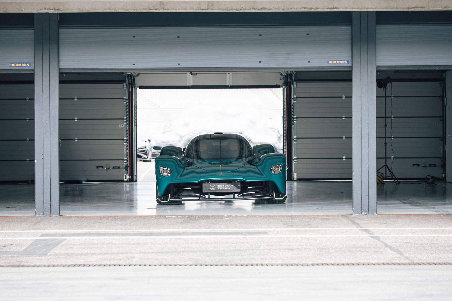 A turquoise Aston Martin Valhalla supercar sits inside a modern garage with its door open.