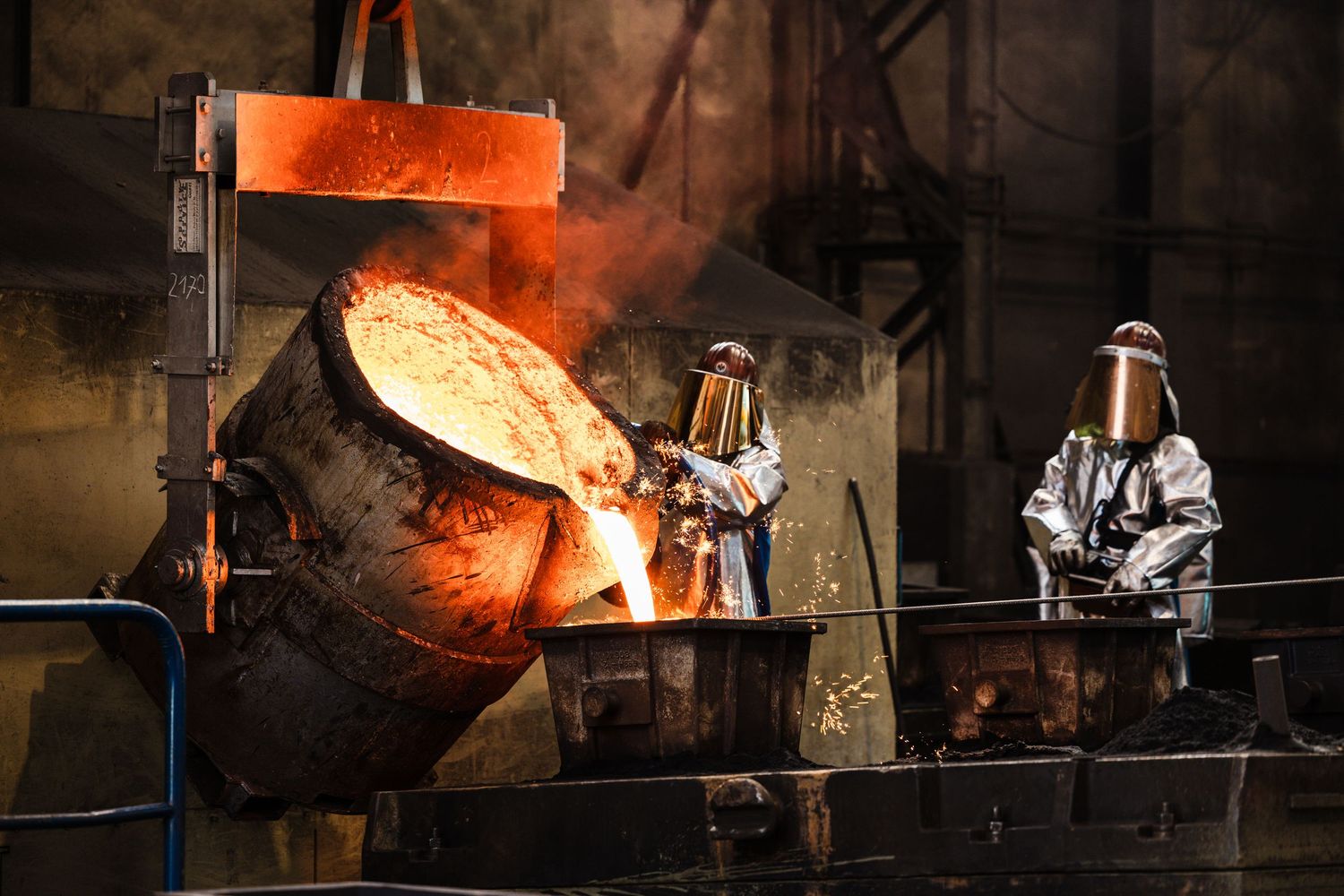 Workers in protective gear observe molten metal being poured from a large industrial crucible in a steel foundry.