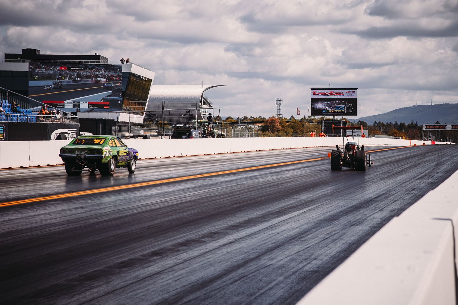 Drag racing vehicles compete on a professional race track under cloudy skies with mountains visible in the distance.
