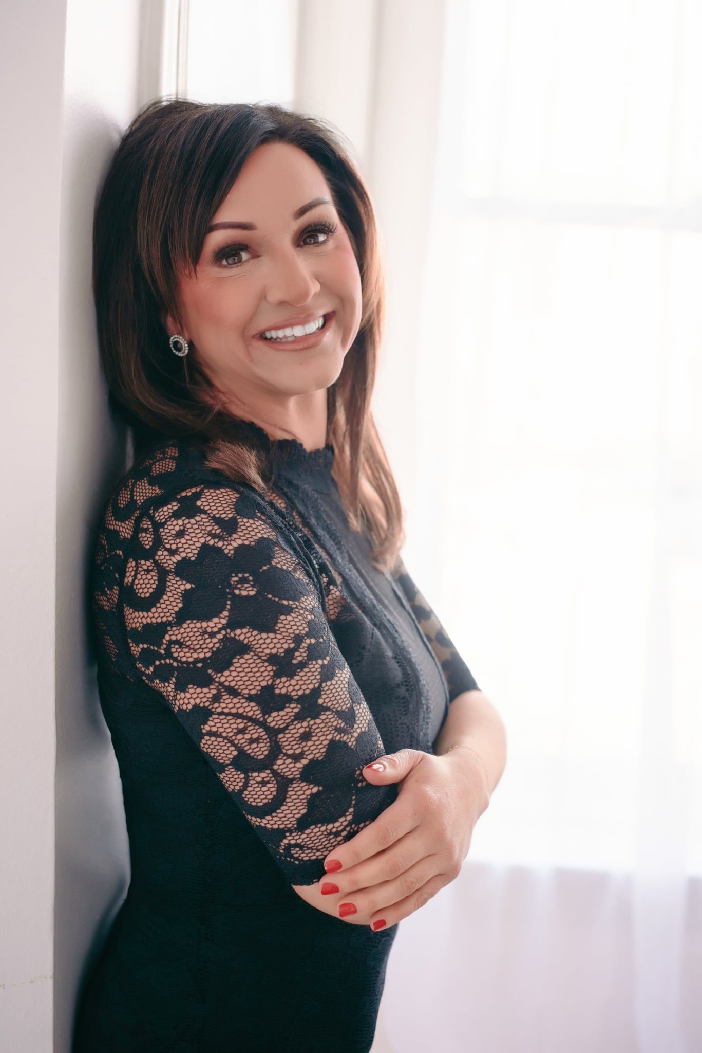 Smiling brunette woman in black lace dress leaning against white wall near window with arms crossed.