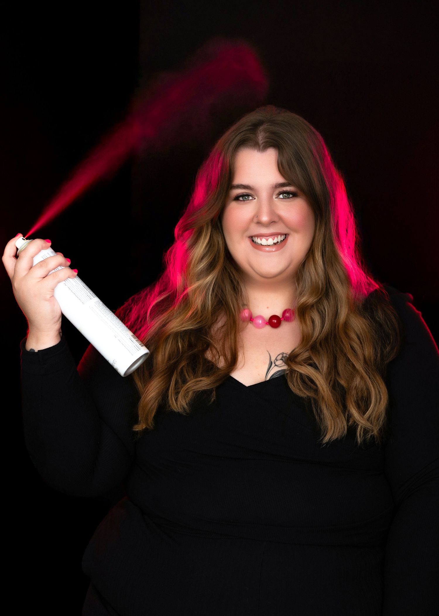Smiling woman in black top holding a white spray can emitting pink smoke, wearing a pink beaded necklace.
