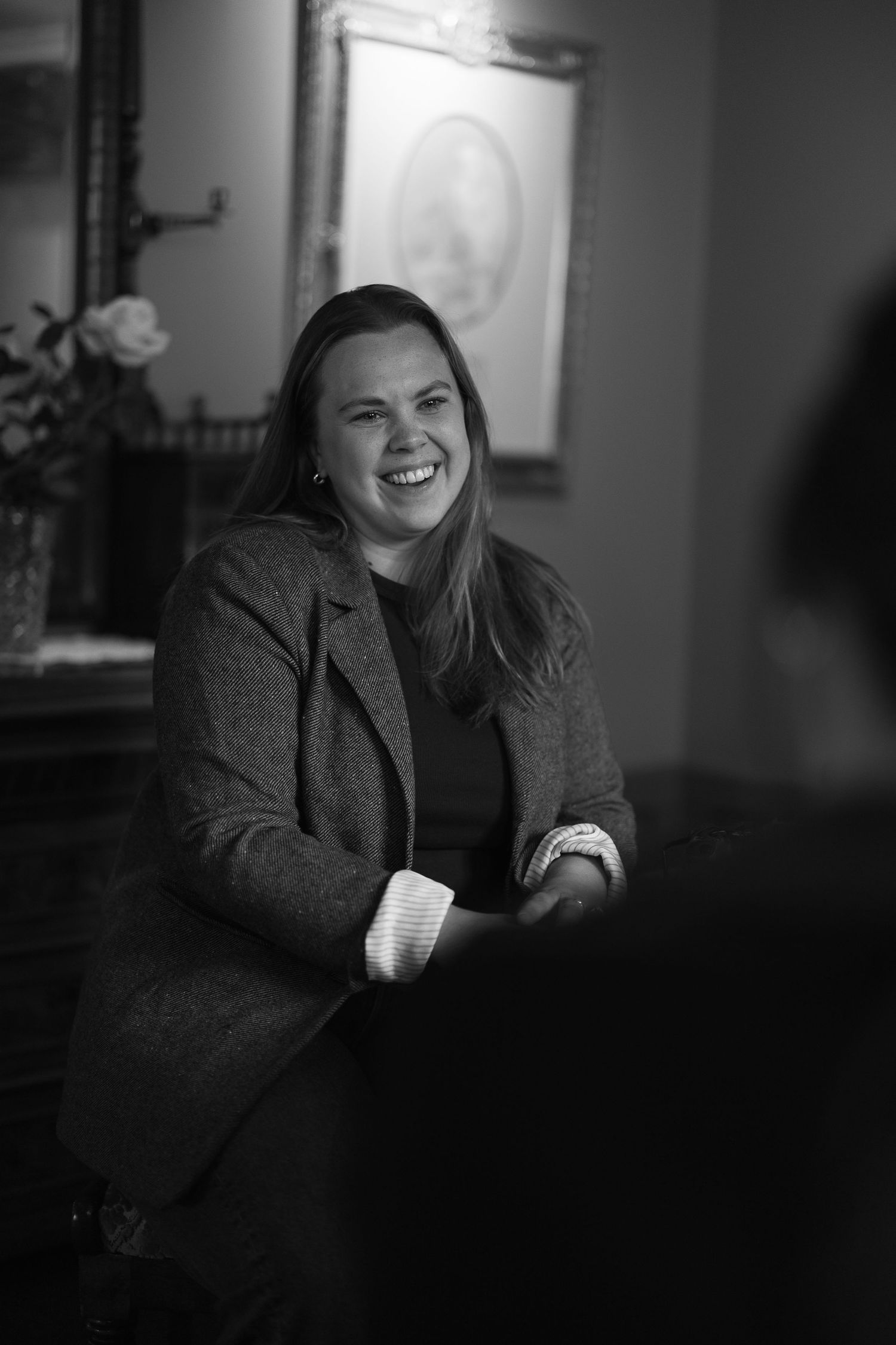 Black and white portrait of a smiling woman with long hair in a tweed blazer seated in a cozy room with framed art.