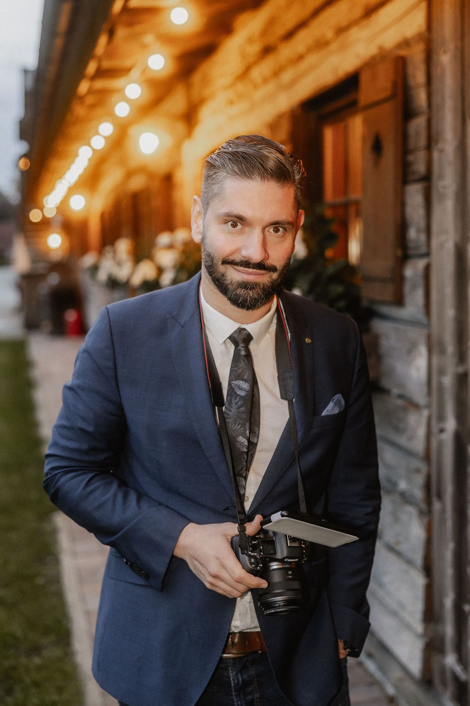 Wedding photographer in navy suit holds camera while posing against rustic venue with string lights.