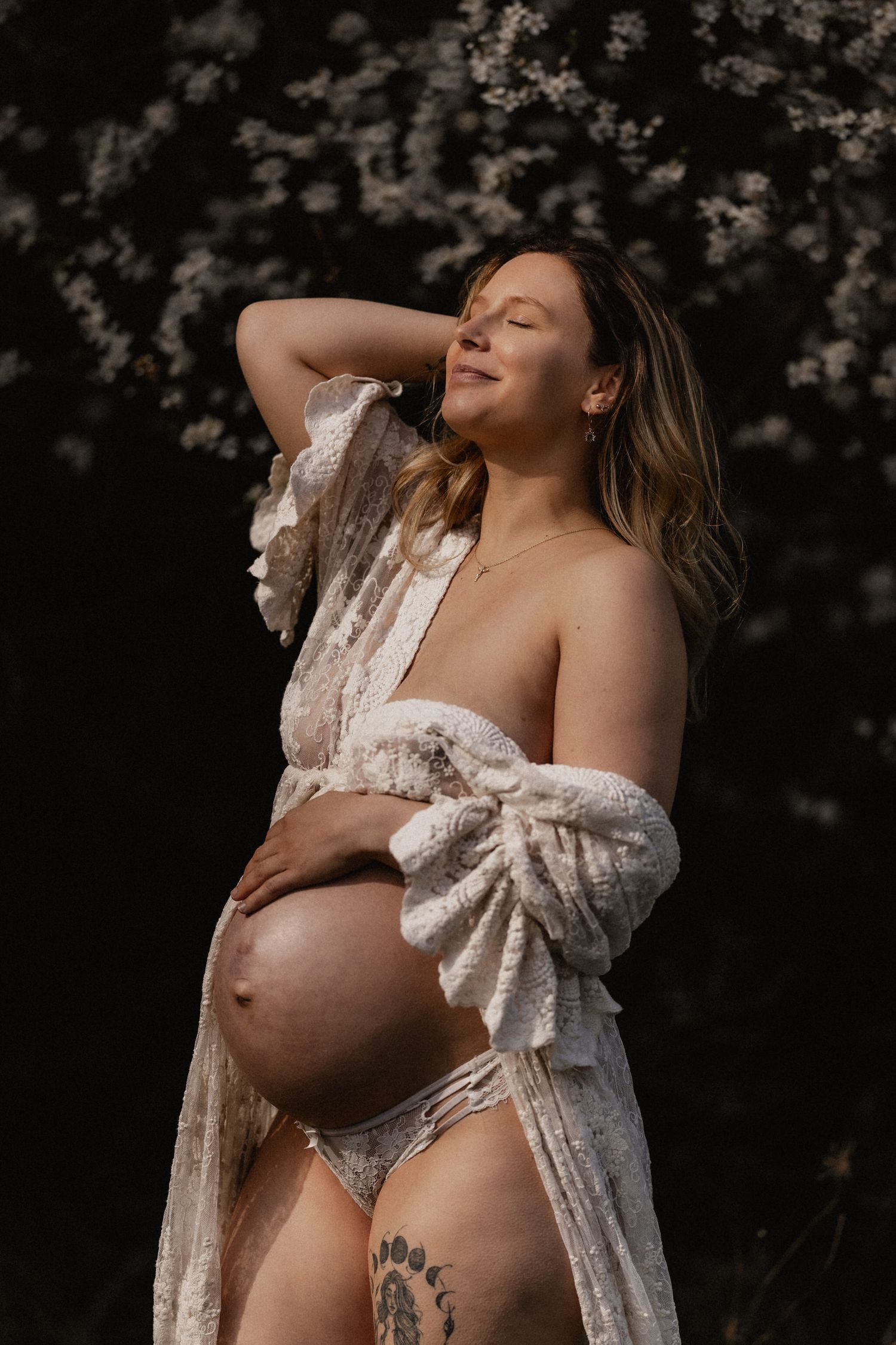 A maternity photoshoot features an expectant mother wearing a white lace robe against a dark background.
