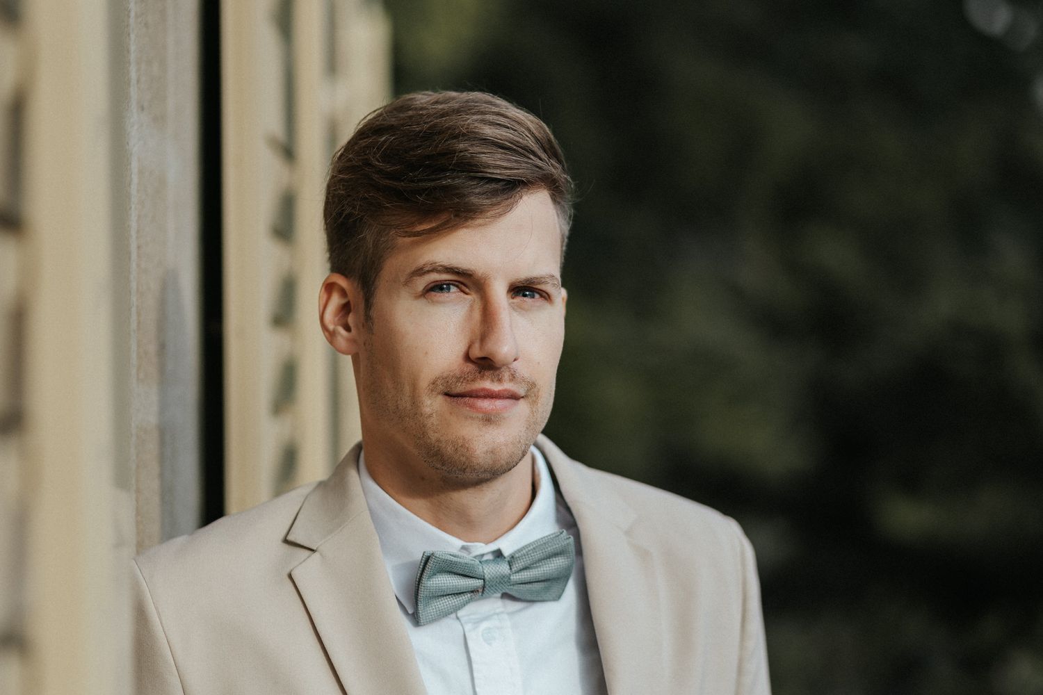A groom in a light beige suit and blue bow tie poses outdoors against a dark background.