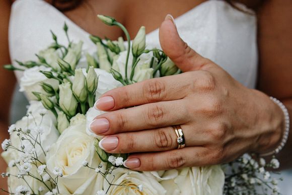 Close-up view of white rose wedding bouquet being held by hands wearing delicate jewelry and white nail polish.