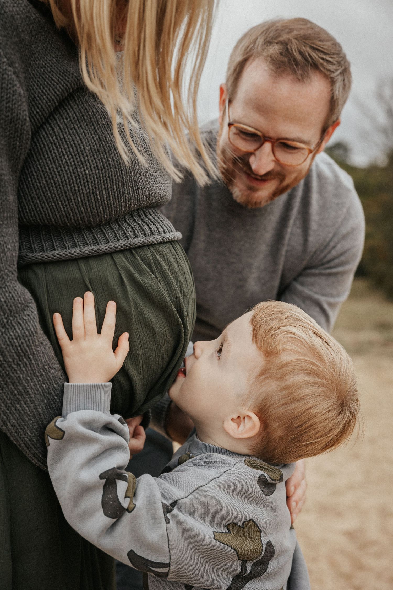 A young boy kisses his pregnant mother's belly while his smiling father looks on outdoors.