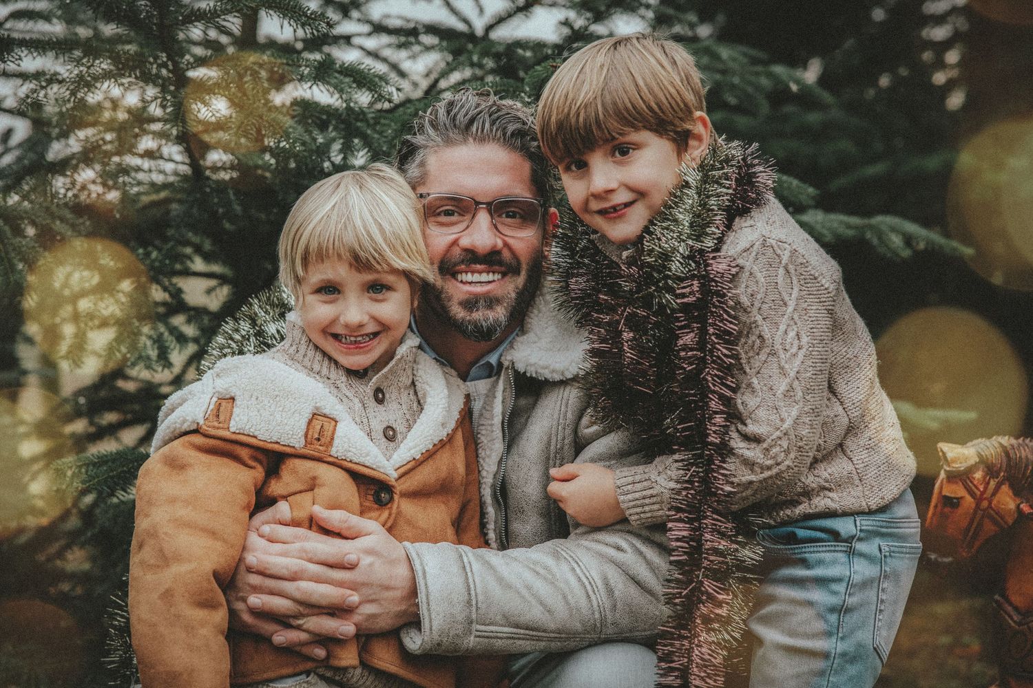 A smiling father hugs his two young children near a Christmas tree with golden ornaments outdoors.