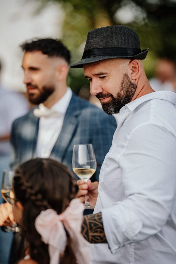 Hochzeitsfotograf Boudier & Koeller – Gast in weißem Hemd und Fedora-Hut hält Glas Weißwein bei Outdoor-Event.