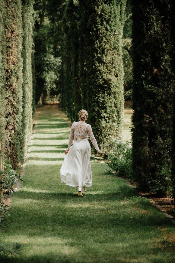 Braut im Spitzenkleid spaziert durch einen von Zypressen gesäumten Gartenweg – verträumt heiraten im Morrhof.