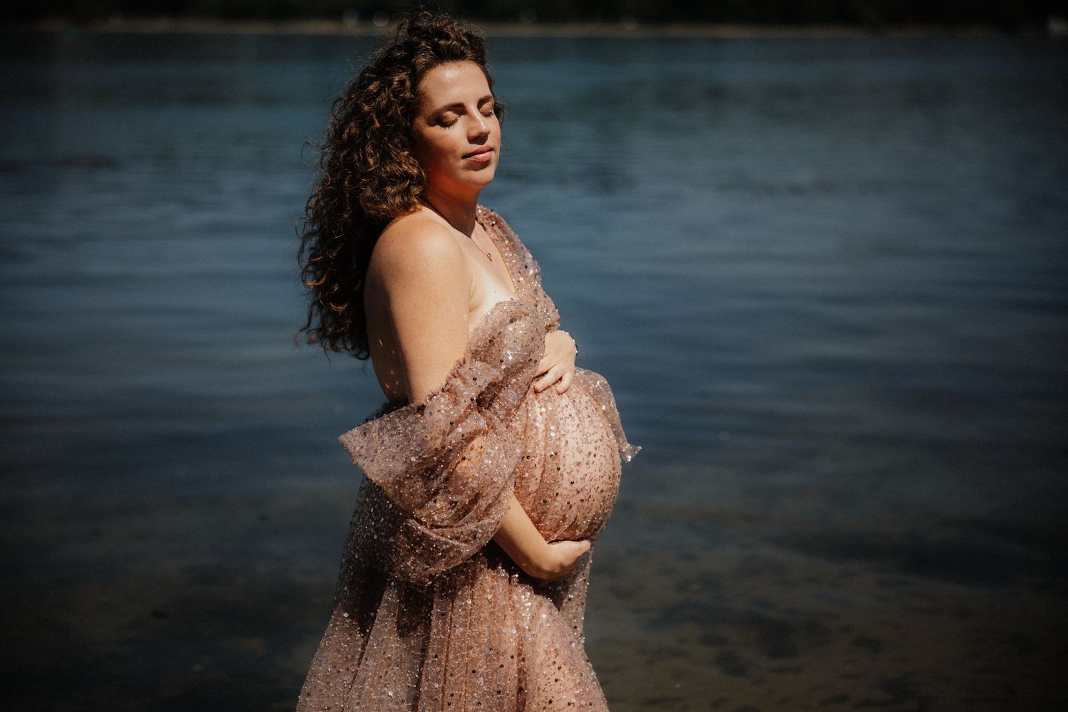 A pregnant woman in a flowing rose gold sequin gown poses gracefully by a serene lake at sunset.