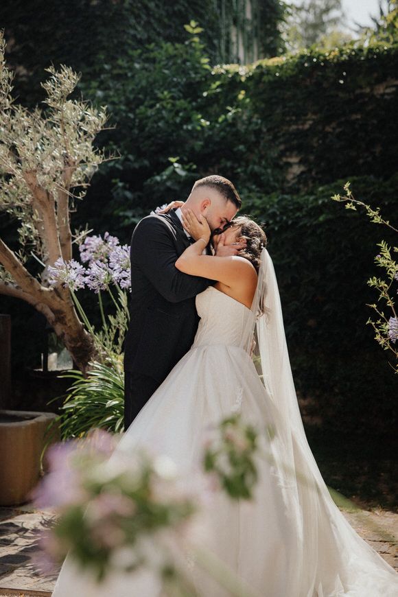 A romantic wedding couple shares an intimate kiss in a garden setting with purple flowers and greenery in the background.