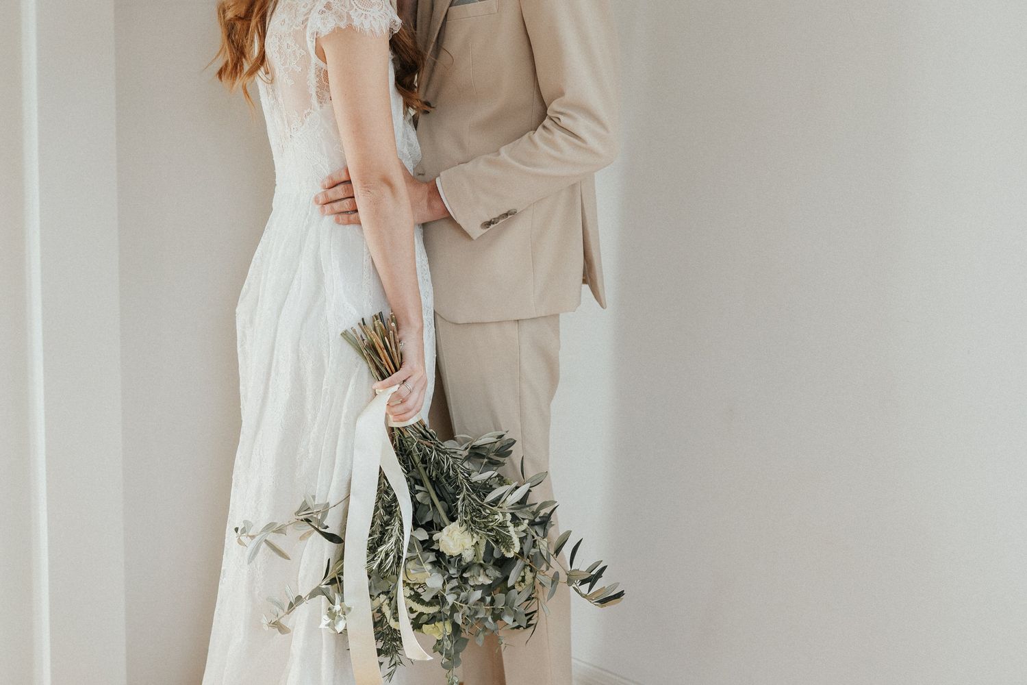 Elegant wedding couple embracing in minimalist white studio with greenery bouquet.