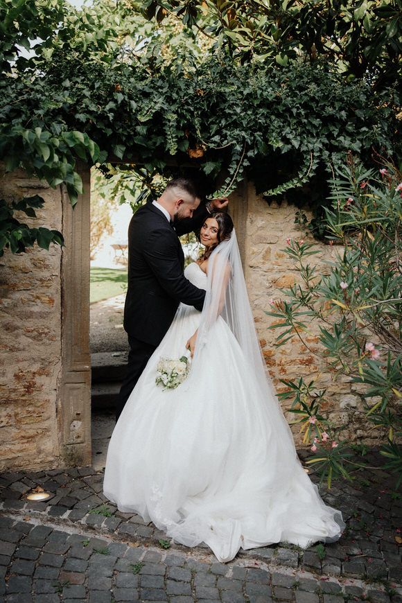 Hochzeitsfotograf Boudier & Koeller – Paar in Hochzeitskleidung teilt intime Momente vor rustikaler Steinwand mit Grün.