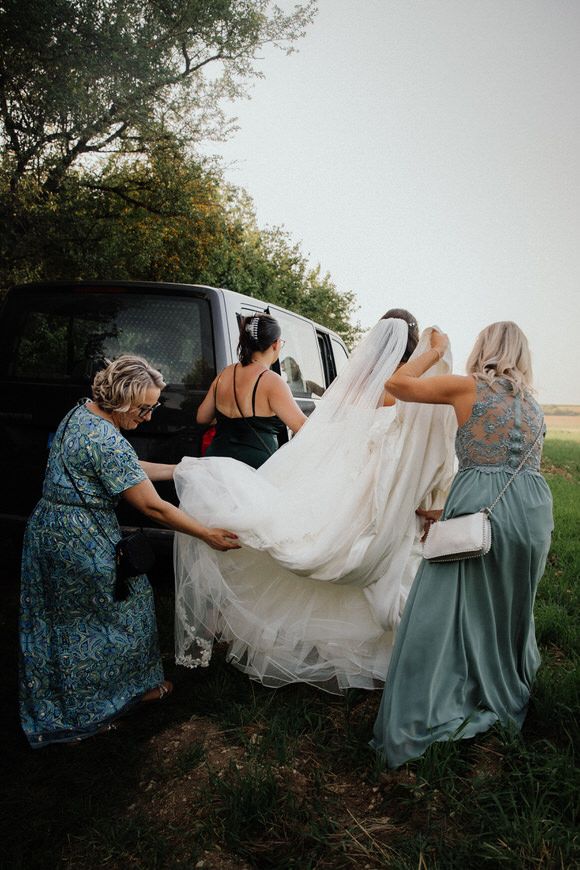 Hochzeitsfotograf Boudier & Koeller – Hochzeitsgesellschaft hilft, Brautkleid bei bewölktem Outdoor-Shooting ins Fahrzeug zu heben.