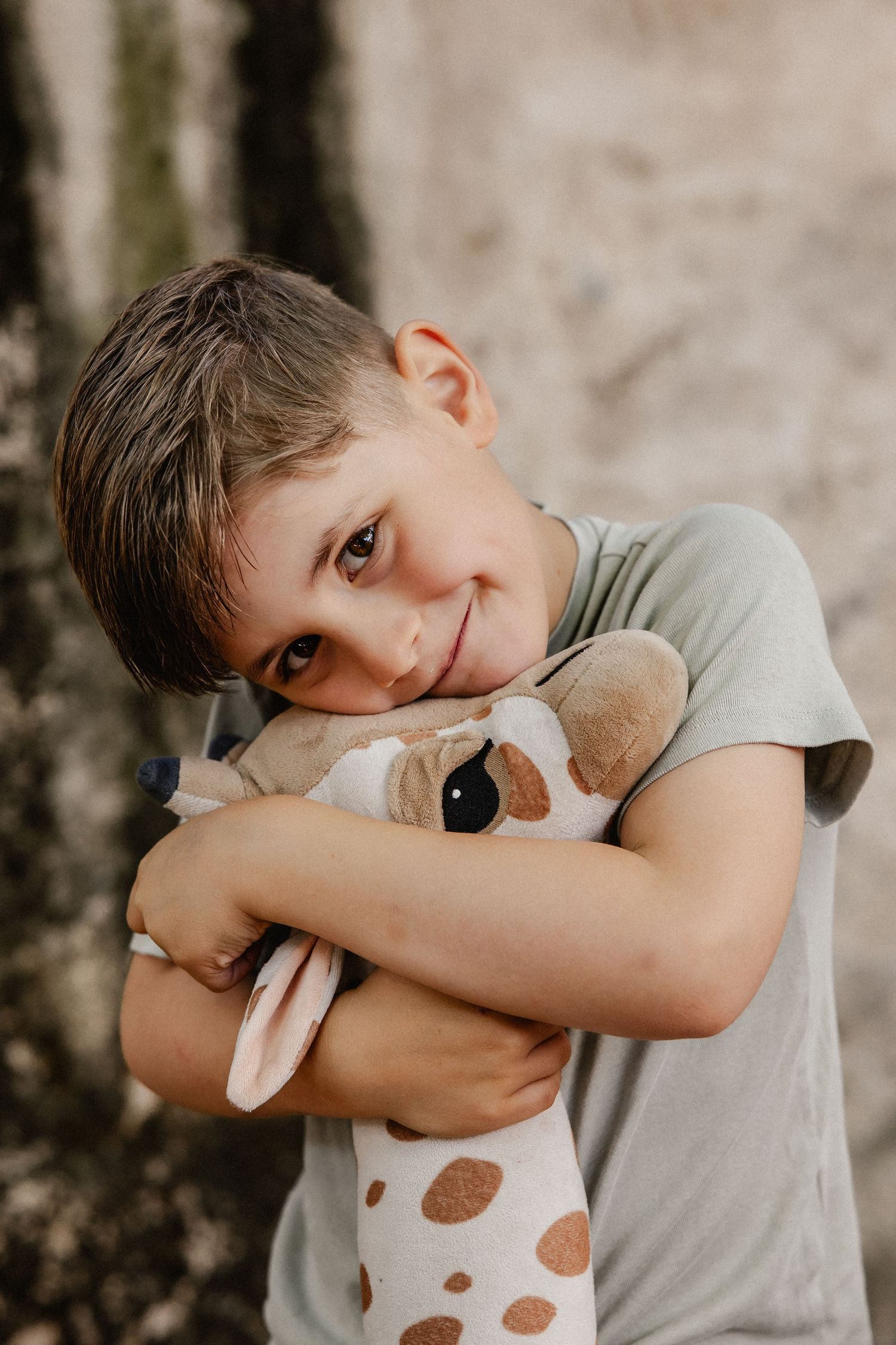 A smiling young boy hugging a giraffe stuffed animal toy outdoors, wearing a gray t-shirt.