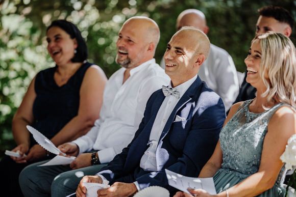 Wedding guests smile and laugh during an outdoor ceremony in a natural garden setting.