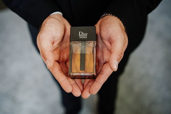 Hands holding a small glass bottle of Dior perfume against a dark background.