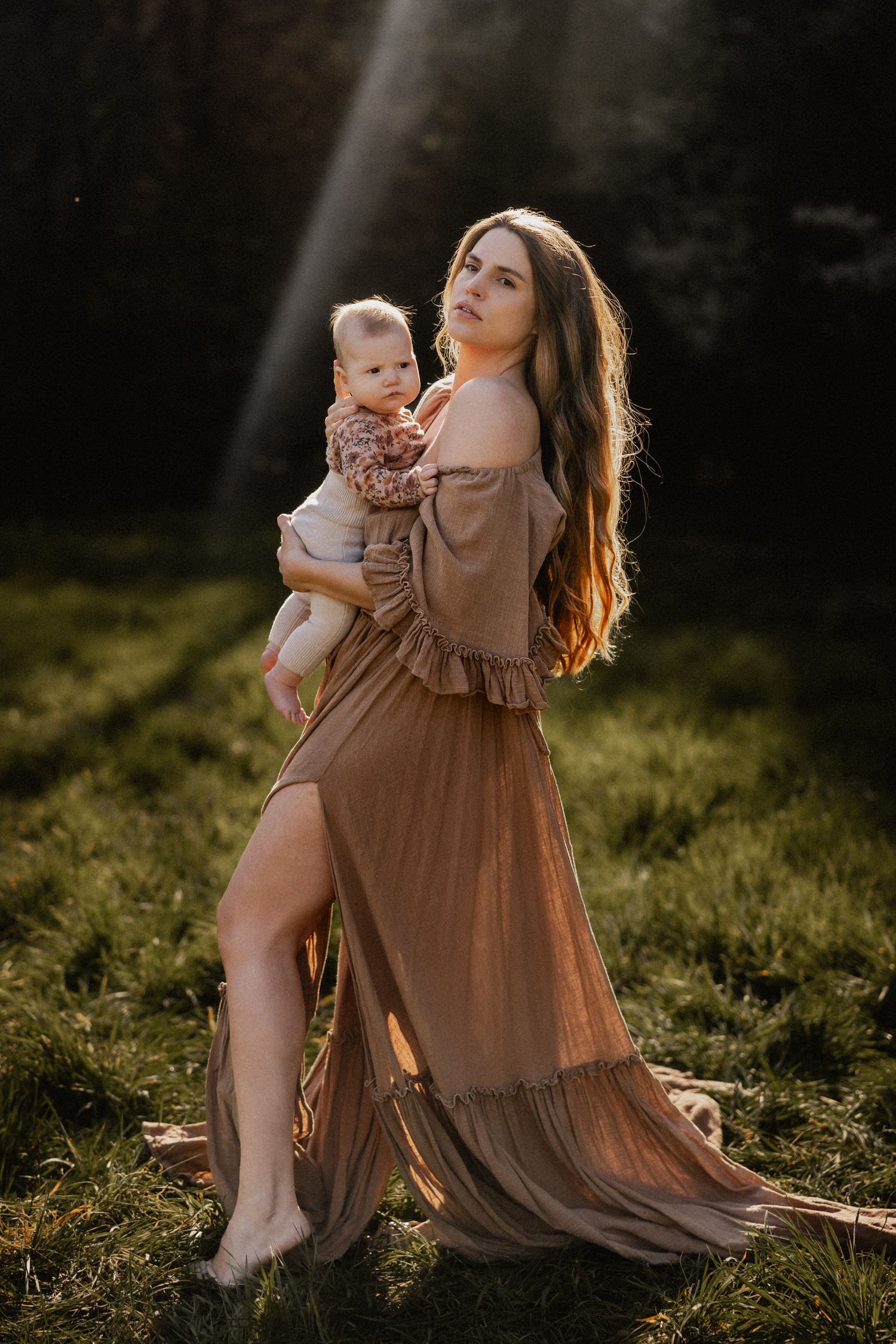 A woman in a flowing brown dress holds a baby while standing in sunlit grass at golden hour.