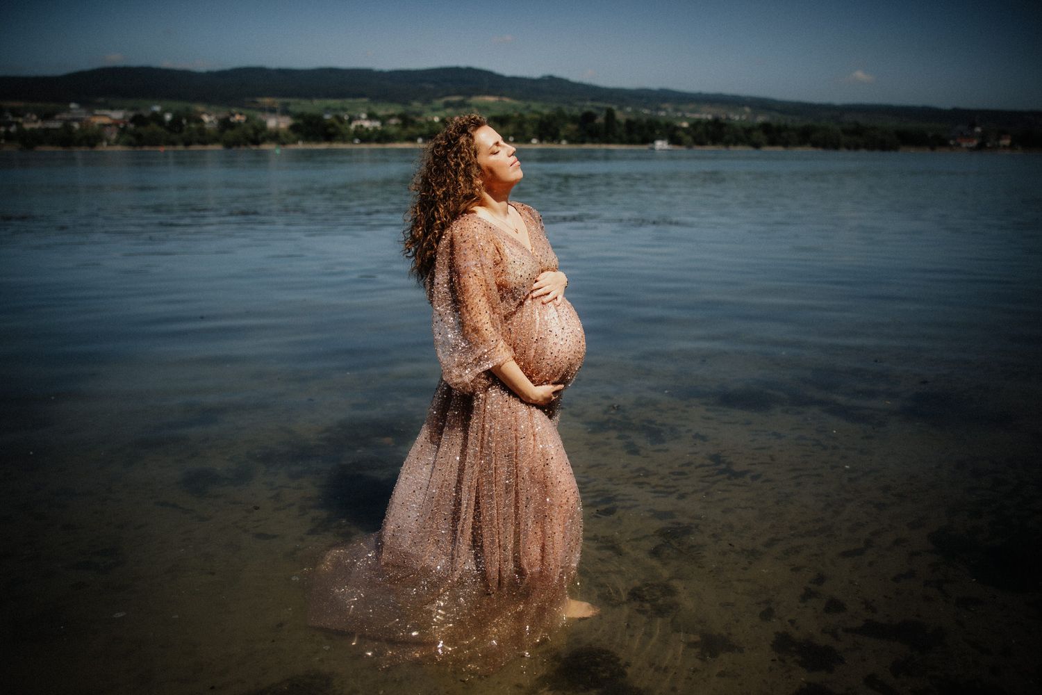 A person in a flowing lace dress stands in shallow lake water at sunset creating a dreamy silhouette effect.