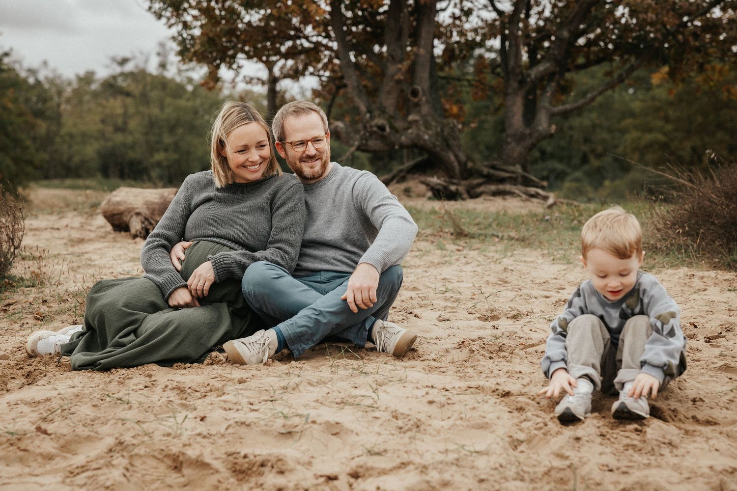 Family of three sitting on sandy ground outdoors, parents smiling with toddler playing in sand near autumn trees.