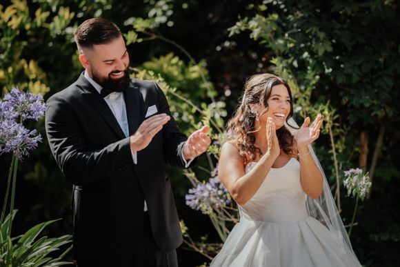 Bride and groom looking at their phones during outdoor garden wedding ceremony with purple flowers in background.
