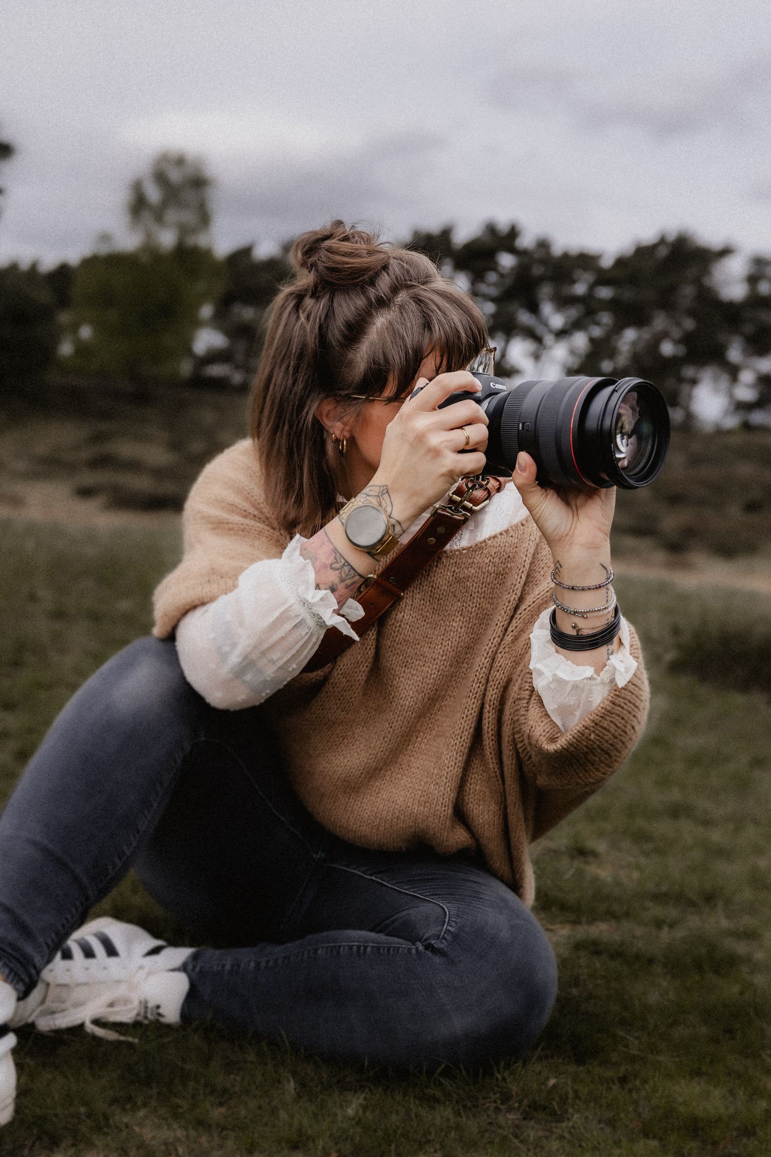 A photographer lying on grass takes photos with a telephoto lens while wearing a brown sweater and jeans.