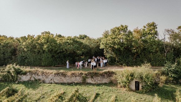 Hochzeitsfotograf Boudier & Koeller – Panoramablick auf rustikale Outdoor-Zeremonie mit Gästen in Holzstühlen.
