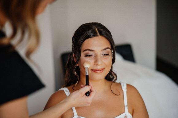 A bride getting her makeup professionally applied while sitting in a white chair during wedding preparations.
