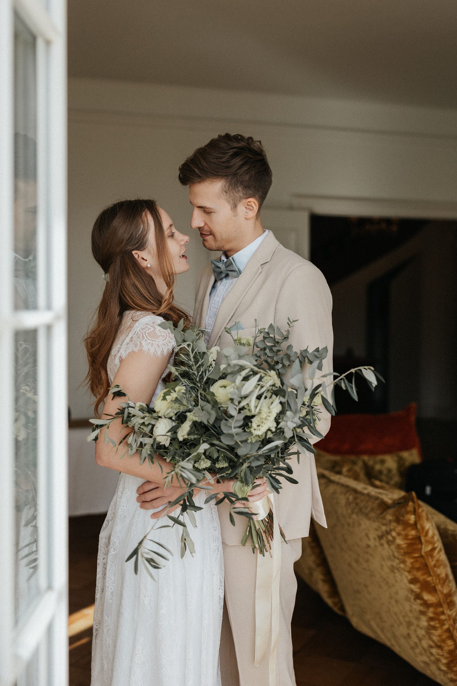 A romantic wedding couple embraces near windows with a beautiful green and white floral bouquet.