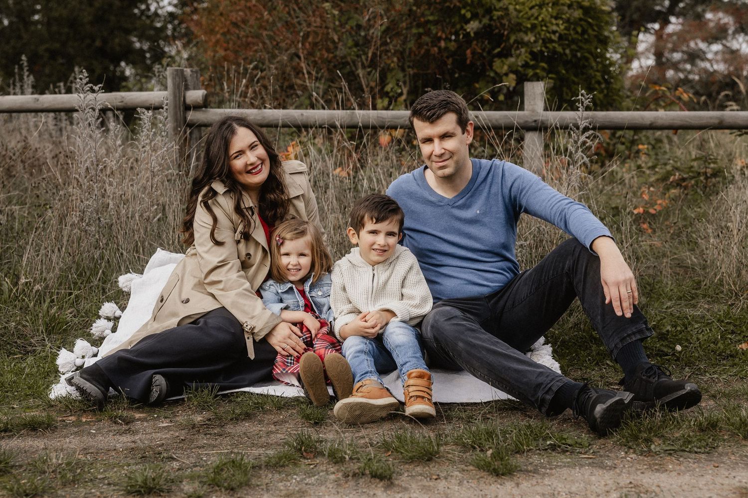 Family of four sitting outdoors on a blanket near a wooden fence, smiling warmly during an autumn photo session.