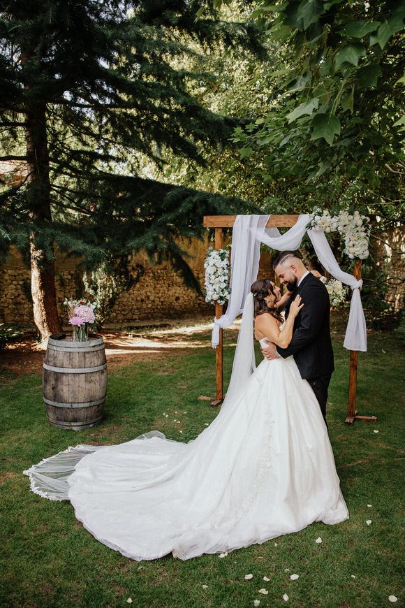 A romantic outdoor wedding scene with a couple dancing on grass near rustic wine barrels and flowing white fabric decor.