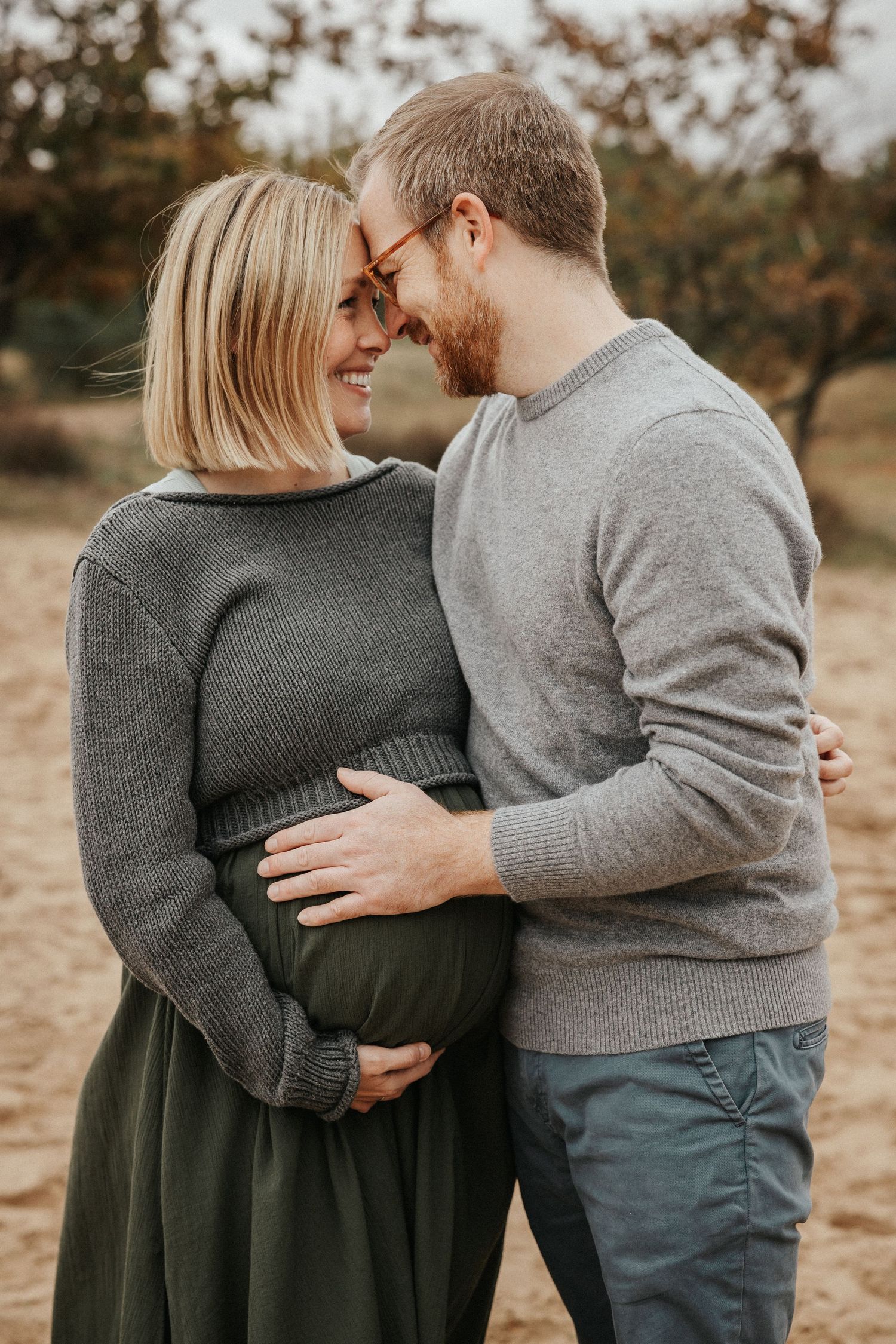 Couple sharing a tender moment outdoors, man embracing pregnant woman's belly, both smiling in cozy gray sweaters.