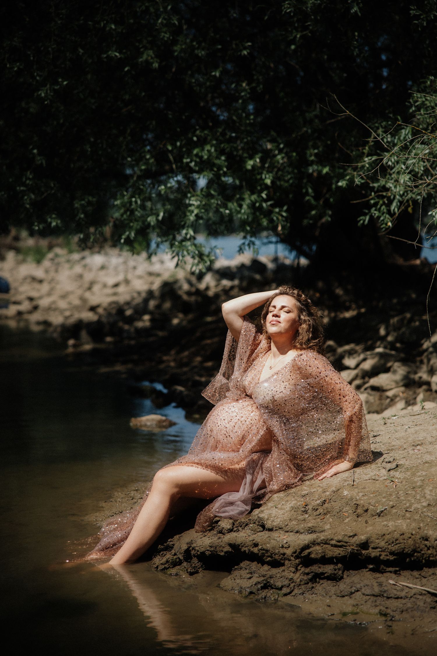 A person in a flowing copper-colored dress poses gracefully on a riverbank in dappled sunlight.