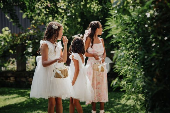 Wedding party members in white and blush dresses gather outdoors in a sunlit garden setting.