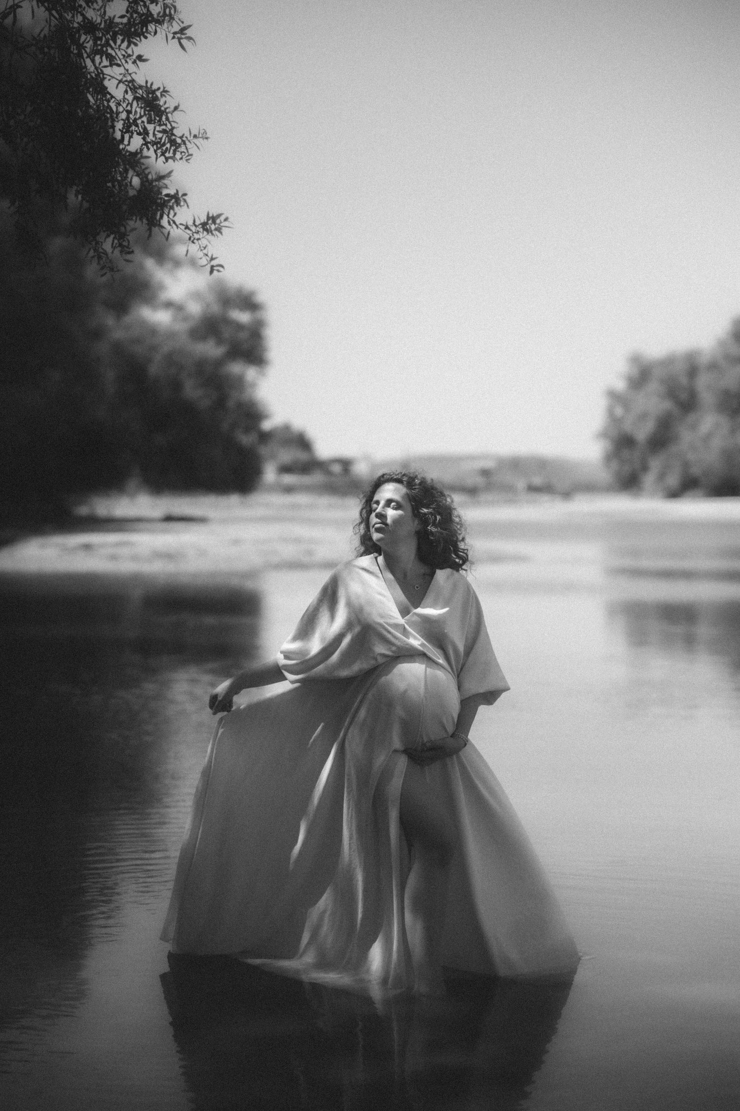 A dreamy black and white portrait of someone in a flowing white dress standing in calm water, surrounded by trees.